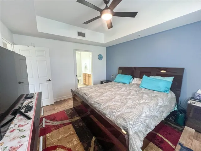 Bedroom featuring a raised ceiling, light wood-style floors, ensuite bathroom, and ceiling fan