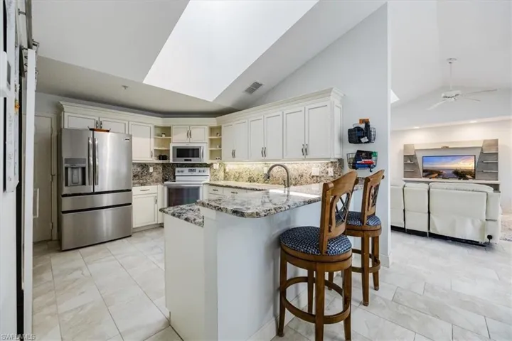 Kitchen featuring white electric range, stainless steel fridge, kitchen peninsula, light stone countertops, and white cabinets