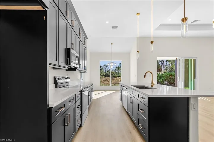 Kitchen featuring stainless steel appliances, light wood-type flooring, a center island with sink, light stone countertops, and dark cabinetry