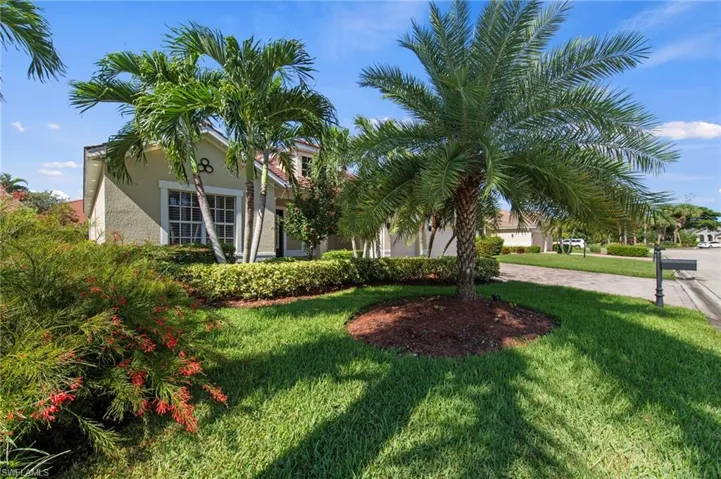 Stucco exterior with a tiled roof, featuring multiple palm trees, a decorative window with grid panes, and a landscaped front yard with lush greenery