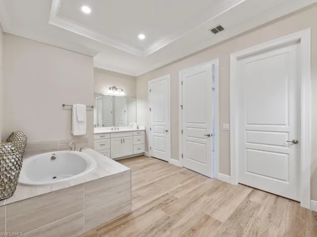 Bathroom with wood-style flooring, a relaxing tiled tub, a tray ceiling, vanity, and crown molding