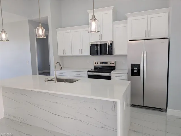 Kitchen featuring stainless steel appliances, light stone counters, white cabinets, decorative light fixtures, and a kitchen island with sink
