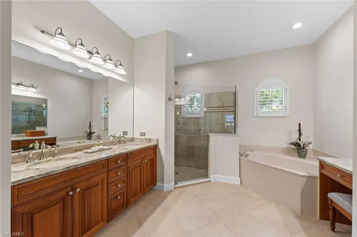 Spacious bathroom featuring an expansive dual vanity with stone countertop and wood-finish cabinetry, a large walk-in shower with glass enclosure and tiled surround, and a built-in soaking tub with decorative arched windows