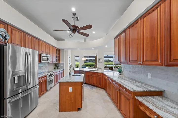 Kitchen featuring wood-finish cabinetry, stainless steel appliances, a center island with sink, light-toned tile flooring, and light-toned countertops