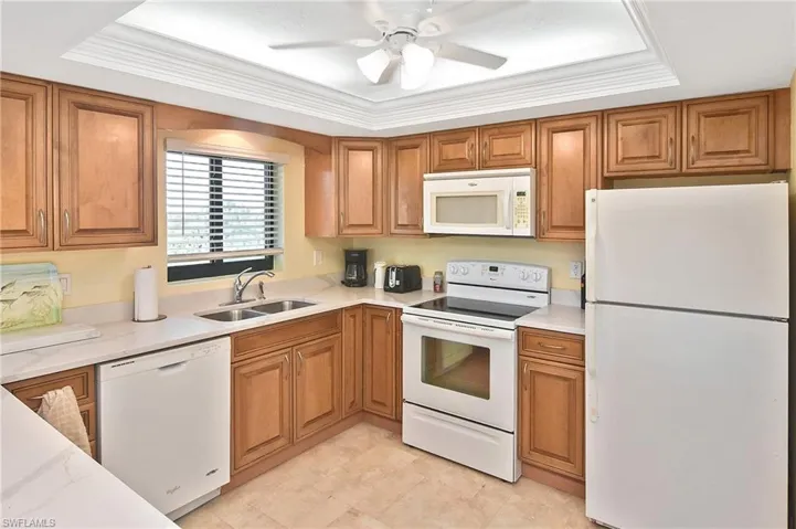 Kitchen with a tray ceiling, white appliances, brown cabinets, light stone countertops, and ornamental molding