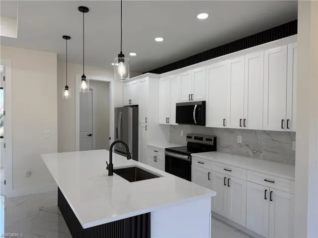 Kitchen featuring stainless steel appliances, decorative backsplash, an island with sink, white cabinetry, and light marble finish flooring