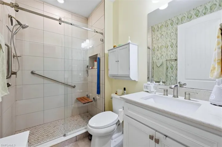 Bathroom featuring a walk-in shower with glass sliding doors, stone-finish tile surround, built-in teak seat, and polished metal hardware, paired with a white vanity featuring a solid surface countertop and undermount sink