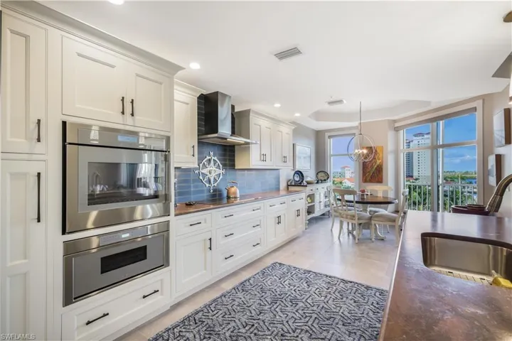 Kitchen featuring wall chimney range hood, stainless steel double oven, backsplash, decorative light fixtures, and white cabinetry