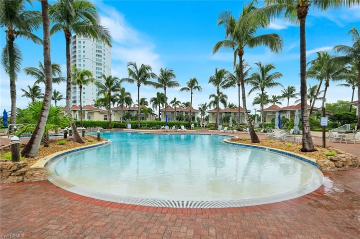 Curved swimming pool with a shallow entry, surrounded by mature palm trees, brick paver decking, and a distant high-rise building