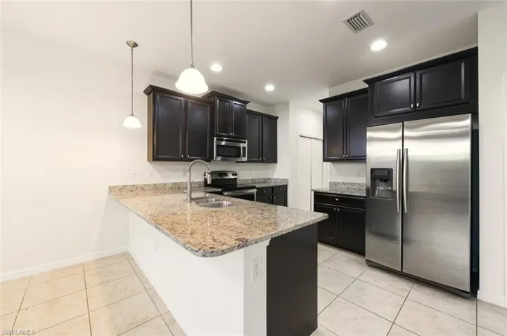 Kitchen featuring appliances with stainless steel finishes, a peninsula, dark cabinetry, light stone countertops, and light tile patterned floors
