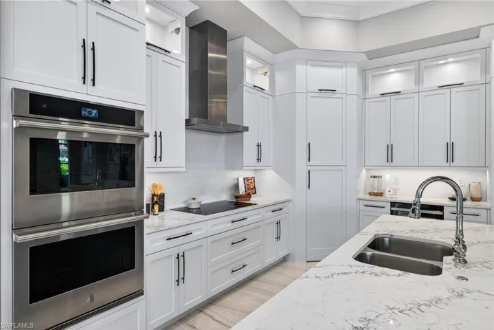 Kitchen featuring stainless steel double oven, glass insert cabinets, light stone countertops, white cabinetry, and wall chimney exhaust hood