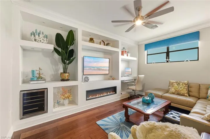 Living area with built in shelves, beverage cooler, ornamental molding, a glass covered fireplace, and wood finished floors