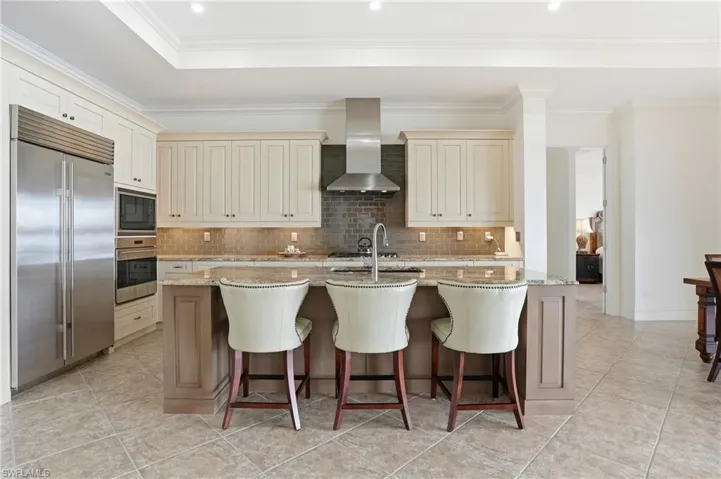 Kitchen with light stone counters, built in appliances, a breakfast bar, ornamental molding, and cream cabinets