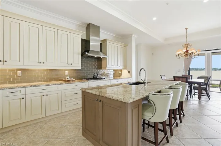 Kitchen with light stone counters, a chandelier, a kitchen island with sink, decorative light fixtures, and wall chimney exhaust hood