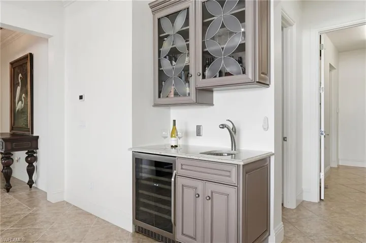 Indoor wet bar with glass insert cabinets, light stone countertops, beverage cooler, light tile patterned floors, and gray cabinetry
