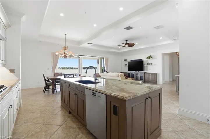 Kitchen featuring decorative light fixtures, a kitchen island with sink, dishwasher, light stone counters, and a raised ceiling