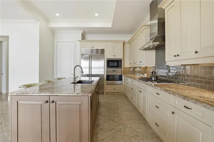 Kitchen with cream cabinets, light stone countertops, built in appliances, decorative backsplash, and wall chimney range hood