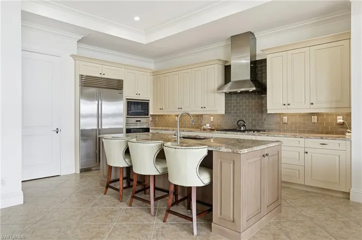 Kitchen featuring cream cabinets, built in appliances, light stone countertops, a breakfast bar area, and wall chimney exhaust hood