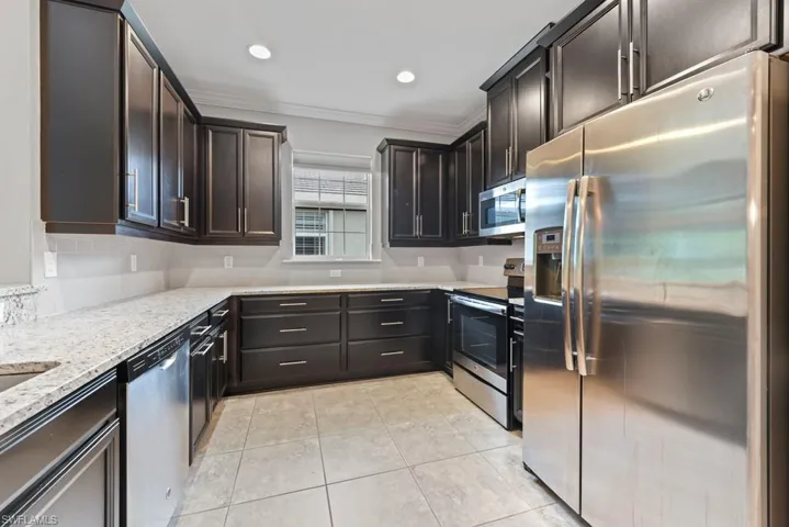 Kitchen with dark brown cabinets, light tile patterned floors, stainless steel appliances, ornamental molding, and light stone counters