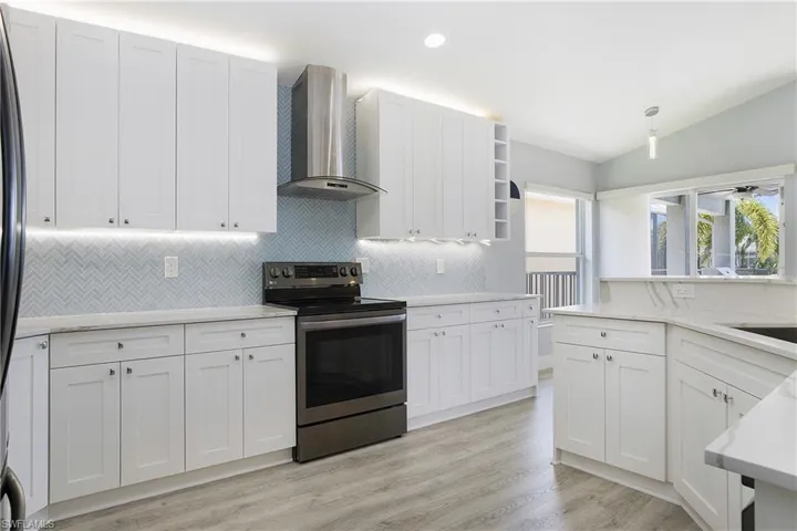 Kitchen featuring stainless steel electric stove, white cabinets, wall chimney range hood, light hardwood / wood-style flooring, and vaulted ceiling