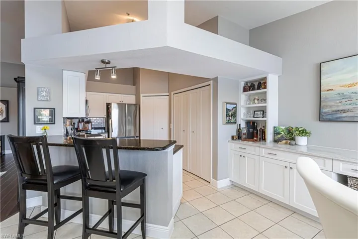 Open-concept living area featuring a kitchen island with a dark countertop, white shaker-style cabinetry, a built-in beverage bar with open shelving, and light-toned tile flooring