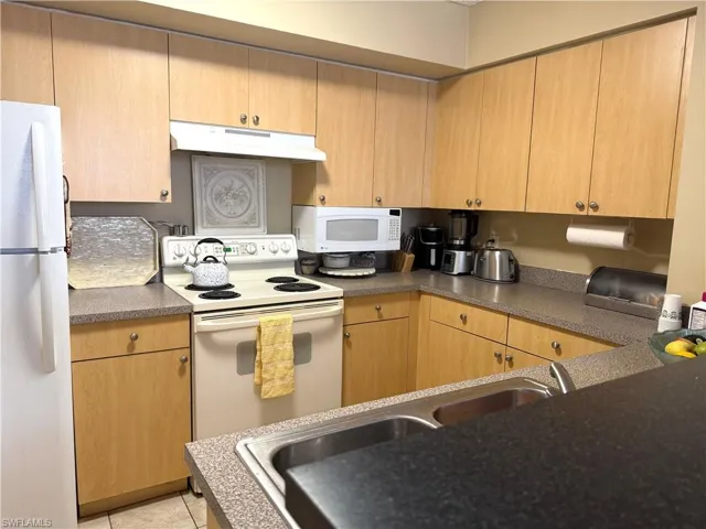 Kitchen featuring a sink, dark countertops, white appliances, under cabinet range hood, and light tile patterned flooring