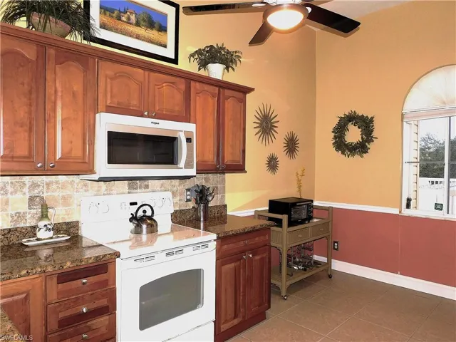 Kitchen featuring white appliances, dark stone counters, decorative backsplash, brown cabinetry, and light tile patterned floors