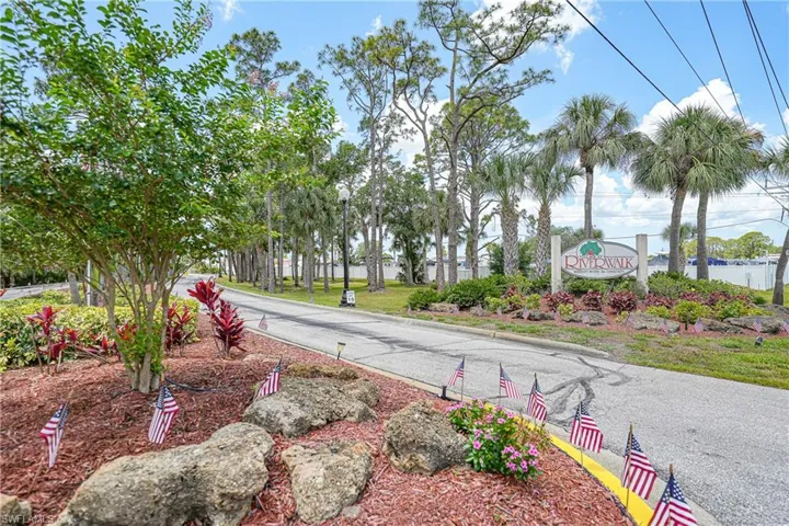 Community entrance featuring a stone sign with "Riverwalk" inscription, mature palm trees, and lush landscaping