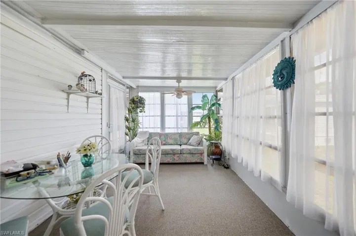 Enclosed sunroom featuring white paneled walls, a neutral carpeted floor, ceiling fan, and multiple windows with sheer white curtains