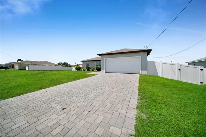 Paver driveway leading to an attached garage, featuring a white vinyl fence and expansive green lawn