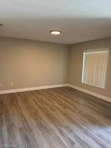 Spacious room featuring wood-finish flooring, neutral-toned walls, a flush-mount ceiling light, and a window with vertical blinds