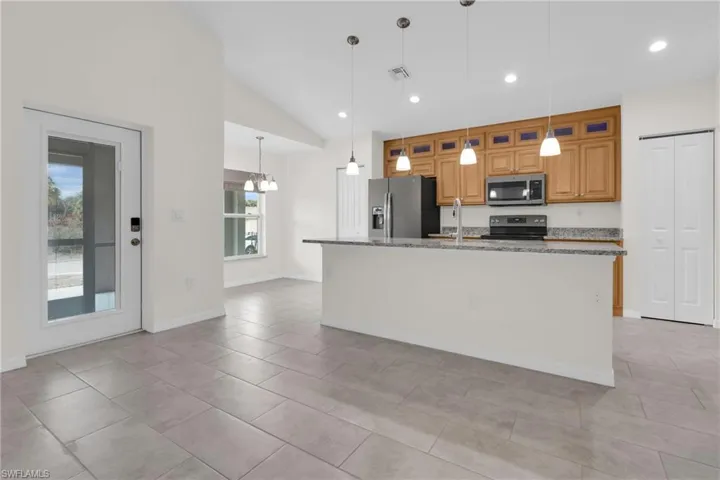 Kitchen featuring vaulted ceiling, wood finish cabinets, light stone counters, stainless steel appliances, and a center island with sink