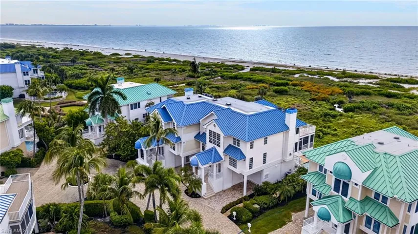Multi-level coastal residence featuring a blue metal roof, elevated architectural design, brick-paver driveway, and exterior chimney, situated along the shoreline