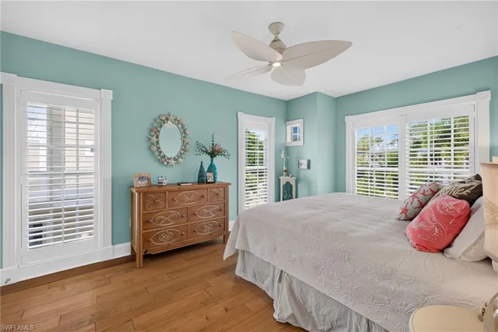 Wood-finish flooring, multi-pane windows with white plantation shutters, and a ceiling fan with leaf-shaped blades