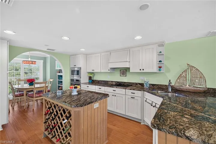 Kitchen featuring white beadboard cabinetry, stone countertops, a central island with built-in wine storage, wood-finish flooring, and a decorative architectural archway