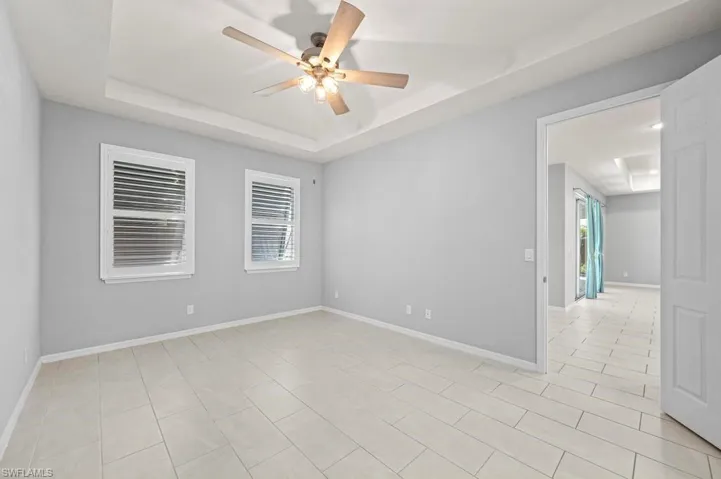 Tray ceiling with a central multi-light ceiling fan, neutral wall tones, ceramic tile flooring, and interior windows featuring white louvered shutters