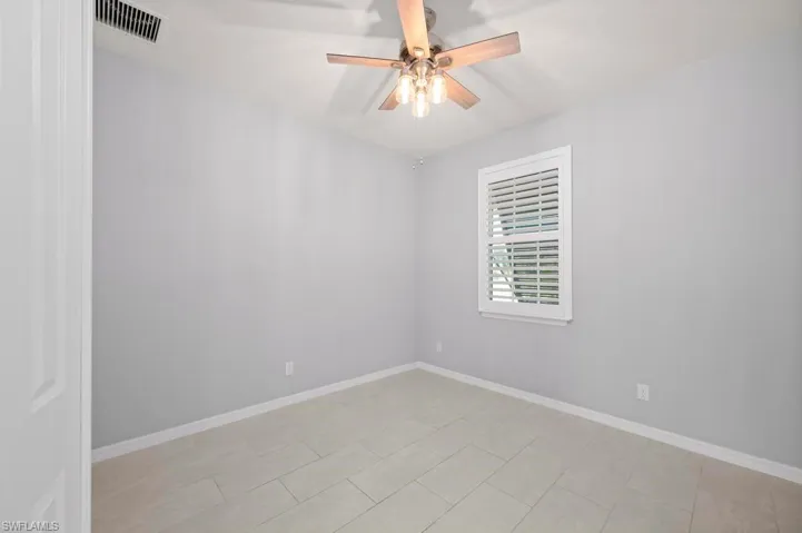 Neutral-toned interior room featuring tile flooring, a multi-blade ceiling fan with a light fixture, and a window with plantation-style shutters