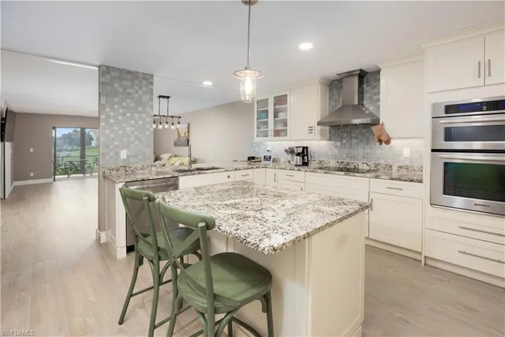 Kitchen with light hardwood / wood-style floors, wall chimney range hood, kitchen peninsula, double oven, and a breakfast bar area