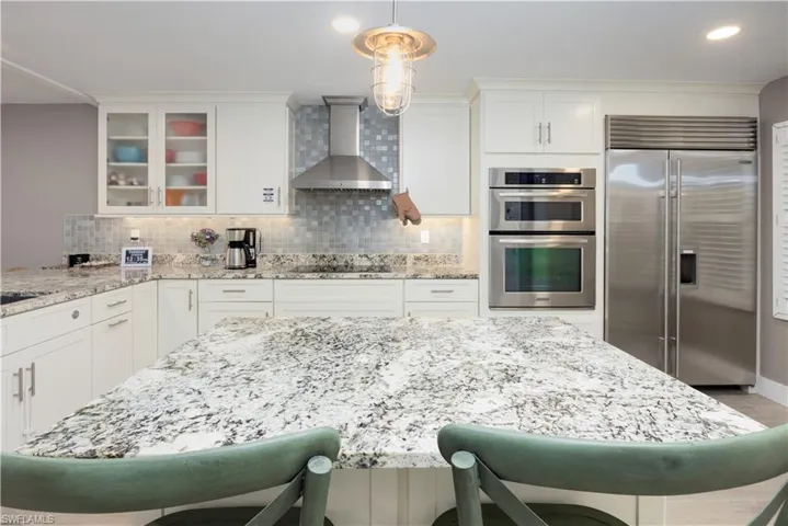 Kitchen featuring backsplash, decorative light fixtures, wall chimney exhaust hood, appliances with stainless steel finishes, and white cabinets