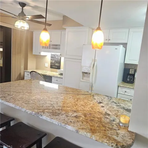 Kitchen with white cabinetry, a kitchen breakfast bar, light stone countertops, white fridge with ice dispenser, and hanging light fixtures