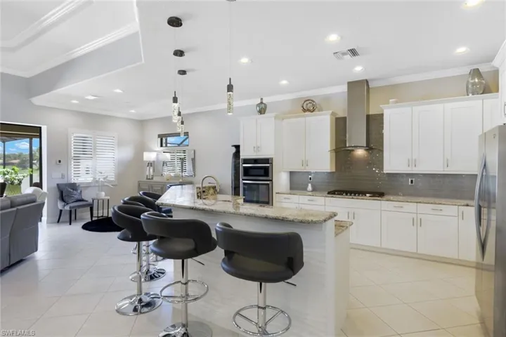 Kitchen featuring stainless steel appliances, a center island with sink, wall chimney range hood, decorative light fixtures, and white cabinetry