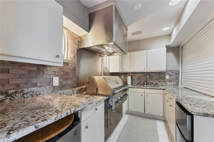 Outdoor kitchen featuring light granite countertops, tasteful backsplash, sink with disposal, wall chimney range hood, light tile floors, and white cabinetry