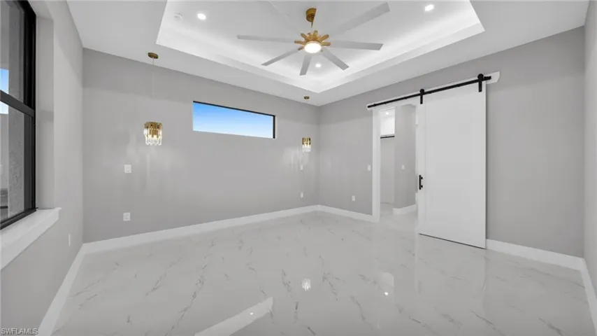Unfurnished bedroom featuring a barn door, ceiling fan, recessed lighting, light marble finish floors, and a raised ceiling