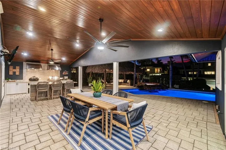 View of patio with plank wood ceiling cladding and modern fans/lighting, Summer Kitchen for grilling, and an outdoor granite island bar with stainless sink and beverage fridge.