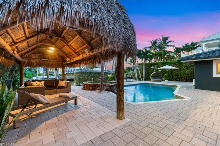 Pool at dusk with a tiki hut, an outdoor living space, an expansive patio area, and an organic custom in-ground pool/hot tub with water feature and “party lights.”