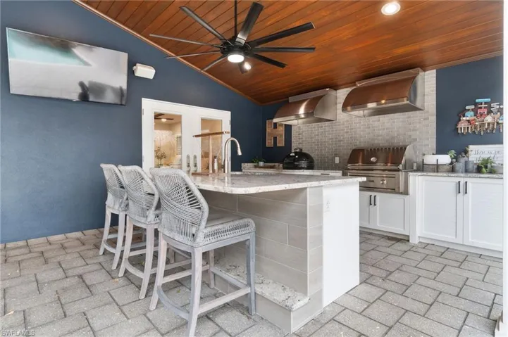 Kitchen featuring white shaker cabinetry, granite counters. wall chimney exhaust hood, wood ceiling, and an island with sink and beverage fridge.