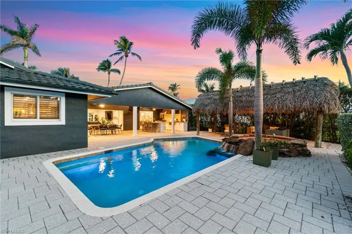 Pool at dusk with a chickee hut, pool waterfall feature, an outdoor granite island bar, and expansive resort style open and covered entertaining patio.