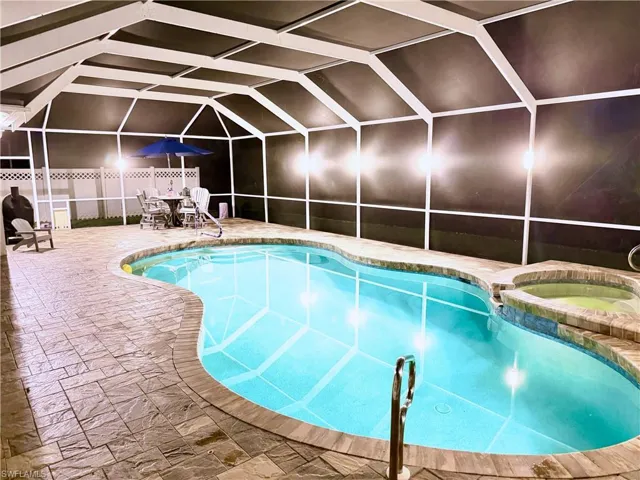 View of pool with bright lights, glass enclosure, a patio area, and a pool with connected hot tub
