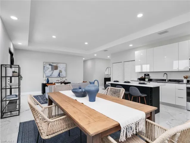 Open-concept living space featuring a dining area with wood-finish flooring, a contemporary kitchen with white gloss cabinetry and a dark island with seating, and recessed lighting throughout