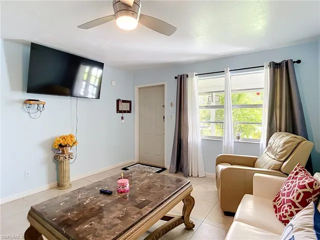 Living space featuring light blue wall paint, tile flooring, a multi-pane window with dark and sheer curtains, a ceiling fan with integrated lighting, and a white entry door